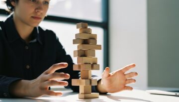 Alt-Text: Eine Person balanciert konzentriert einen Stapel unregelmäßiger Holzklötze auf einem Schreibtisch, um Projektrisiken zu minimieren.. Editorial Style Fotografie, aufgenommen mit einer 35mm-Kamera, die eine Person in einem modernen, minimalistischen Büro zeigt. Die Person, deren Gesicht konzentriert, aber nicht gestresst ist, balanciert sorgfältig einen Turm aus verschieden geformten Holzklötzen. Das Licht fällt durch ein Fenster und erzeugt weiche Schatten. Der Hintergrund ist unscharf (Bokeh), der Fokus liegt auf den Händen und den Klötzen. Kodak Portra 400 Stil, leichte Filmkörnung, natürliche Hauttextur sichtbar, gedämpfte, naturgetreue Farben. Medium Shot.