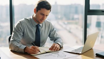 Berater plant eine Google Ads Kampagne zur Lead-Generierung in seinem Büro. | Prompt: Editorial style medium shot, high resolution photography. A sharp, professionally dressed male consultant in his late 30s sits at a large wooden desk in a bright, modern office with a blurred background (bokeh). He is not looking at a laptop, but thoughtfully sketching a funnel diagram in a high-quality notebook with a fountain pen. The morning light streams in from a large window, creating a sense of clarity and focus. The atmosphere is one of calm, strategic planning and expertise. Shot on a 35mm camera, Kodak Portra 400 style, true-to-life colors, slight film grain, visible skin texture.